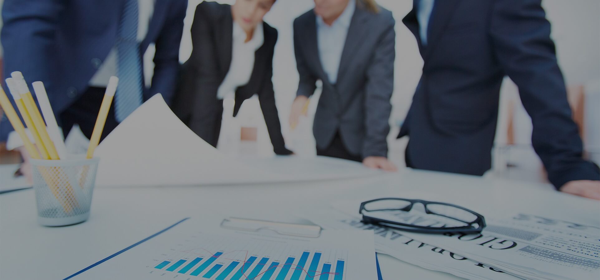 4 business people standing at a work desk with the focus on a container of pencils, a spreadsheet, reading glasses and a newspaper